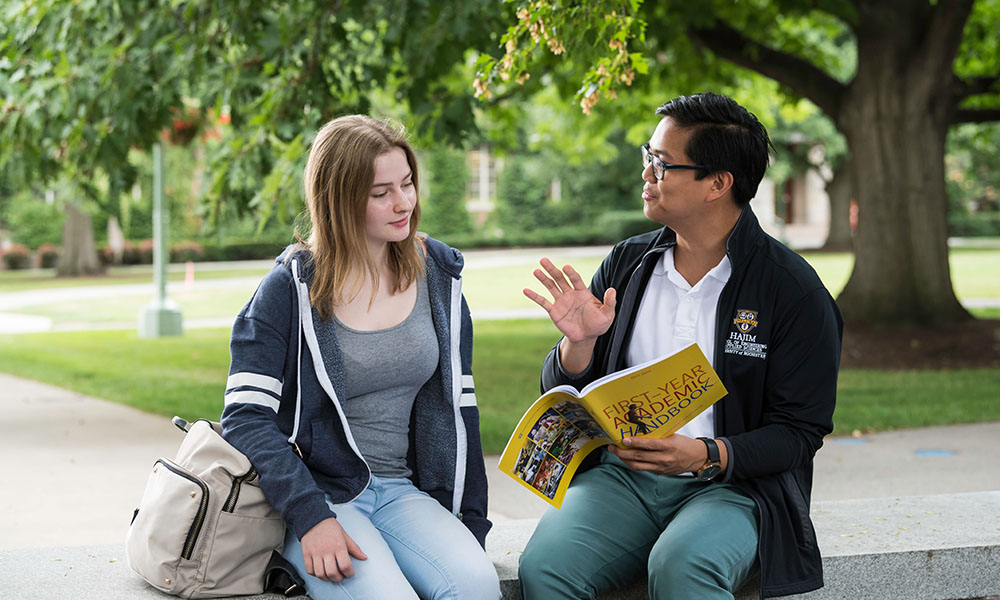 A student and advisor meeting outdoors on a bench to review a catalog.
