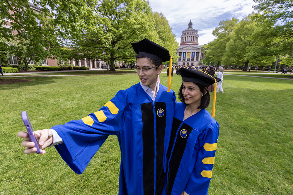 Two students in graduation caps and gowns taking a selfie on Eastman Quad.