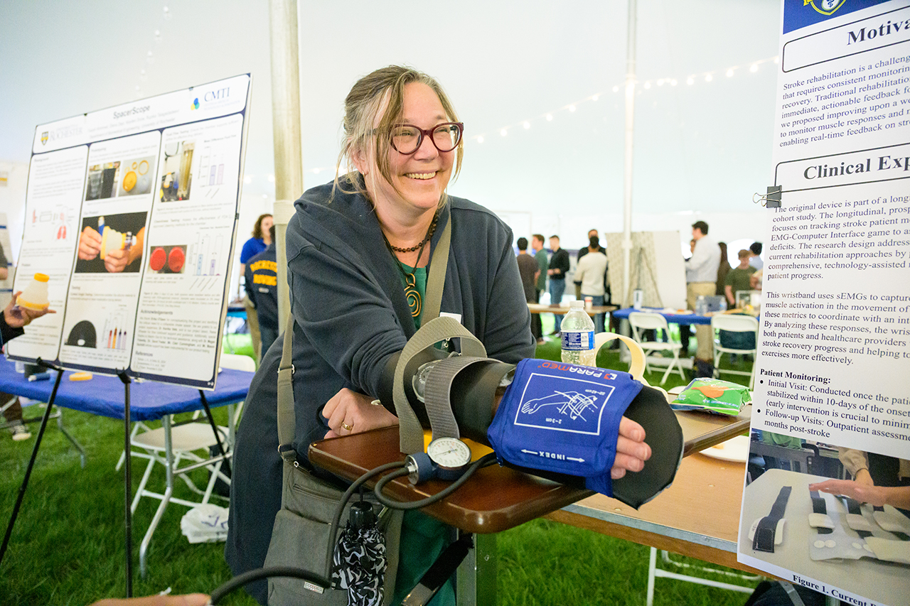 A smiling person putting their forearm into a medical cuff at a poster presentation.