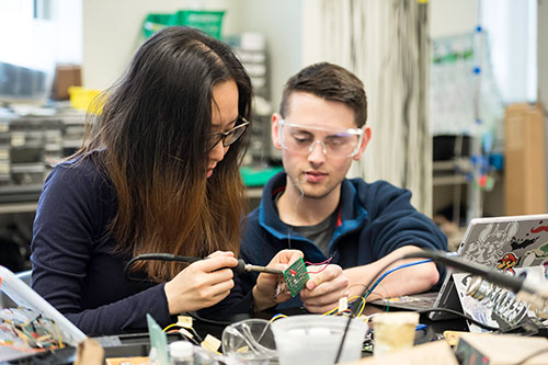 An undergraduate student soldering will another student looks on.