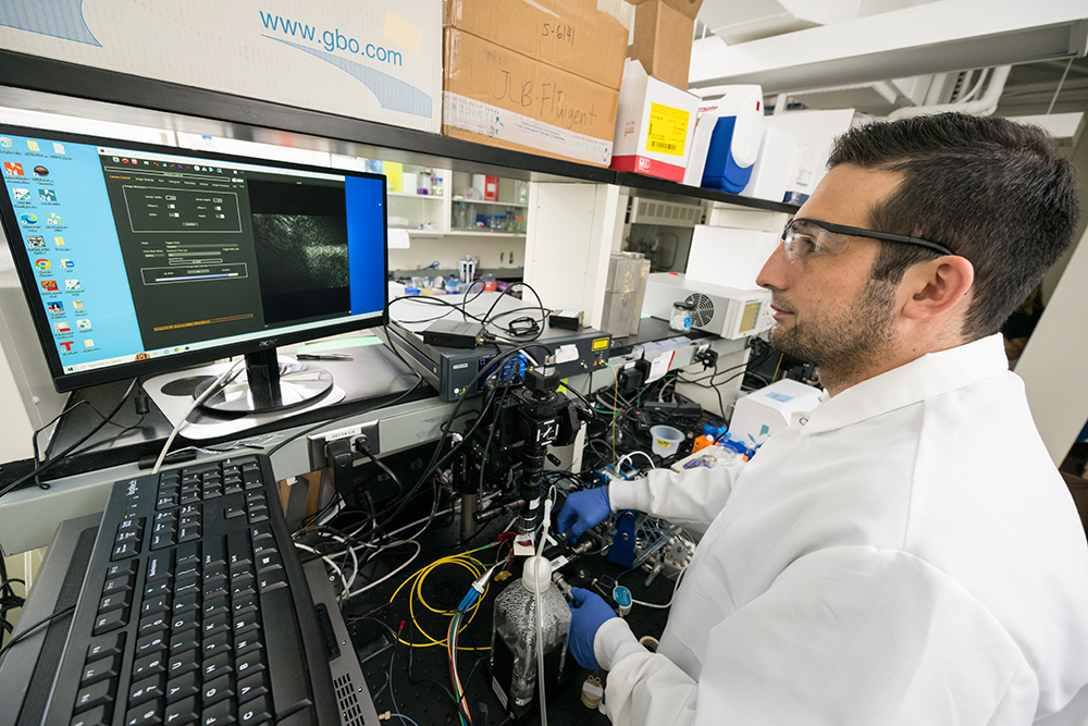 A graduate student in a lab coat and safety glasses uses equipment to take measurements and view them on a computer screen.