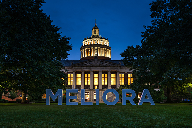 Large white letters spelling the word Meliora on the Eastman Quad at night with Rush Rhees Library lit up in the background.