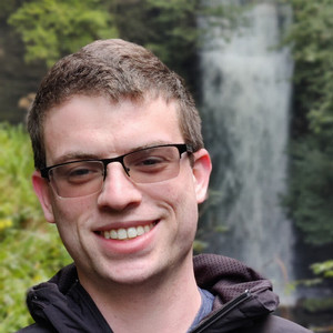 Man smiling at camera wearing glasses while outside by a tree.