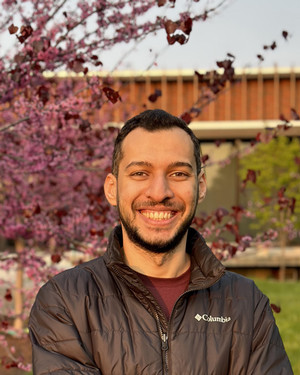 Eslam smiling at camera in outside setting with pink blooming tree in background