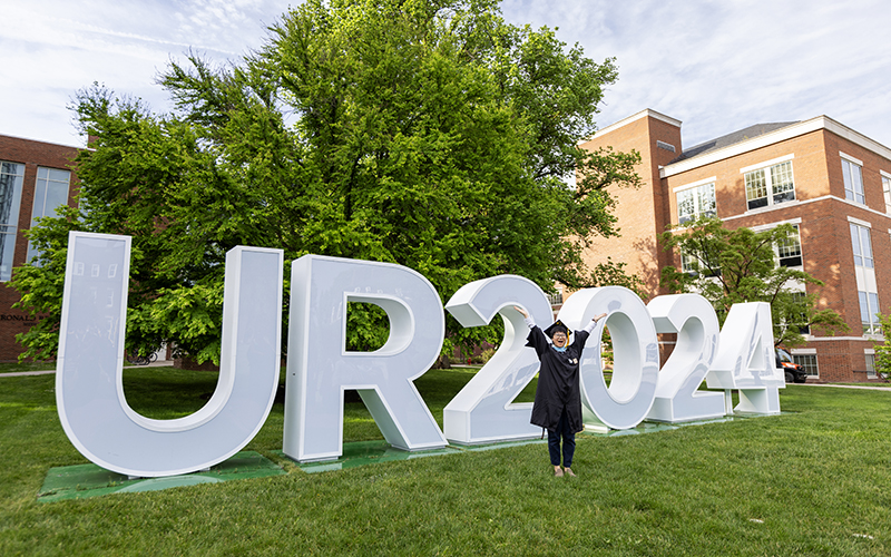 Large letters spelling UR2024 with a graduate standing in front of them.