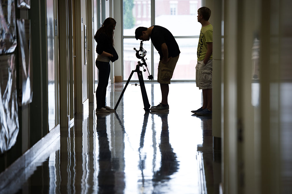 Three students in silohuette standing in a hallway with a telescope pointed towards a large window.