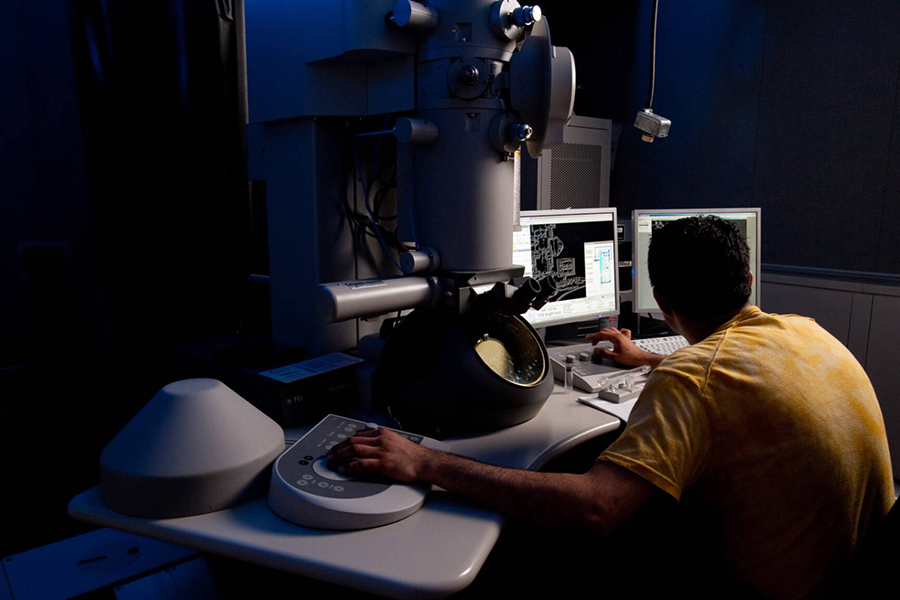 A person in a yellow shirt seen from behind works with a transmission electron microscope in a darkened room.