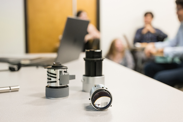 Three lenses in the foreground sitting on a table with a laptop and some students blurred in the background.