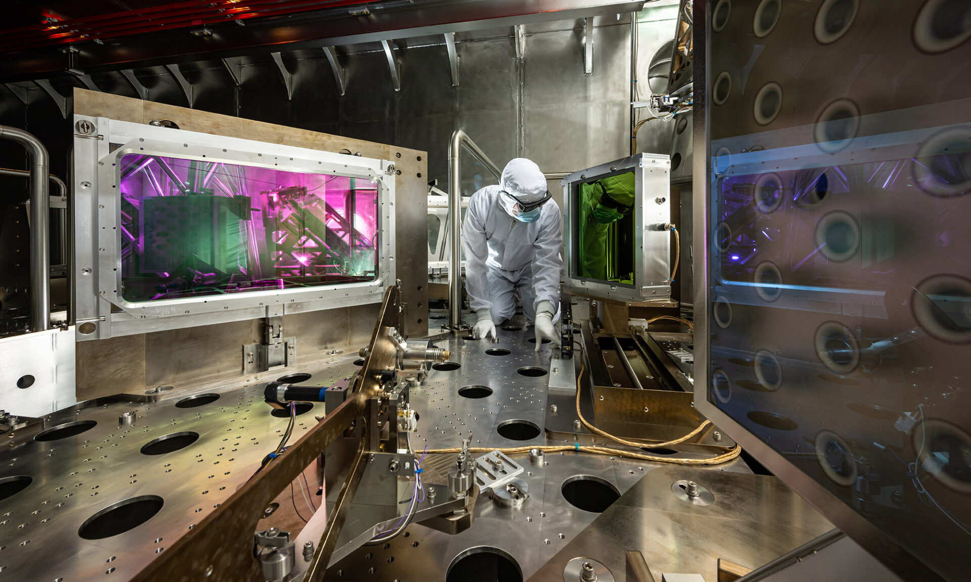 Person in protective gear works inside a high-tech laser research facility with glowing panels and precision equipment.