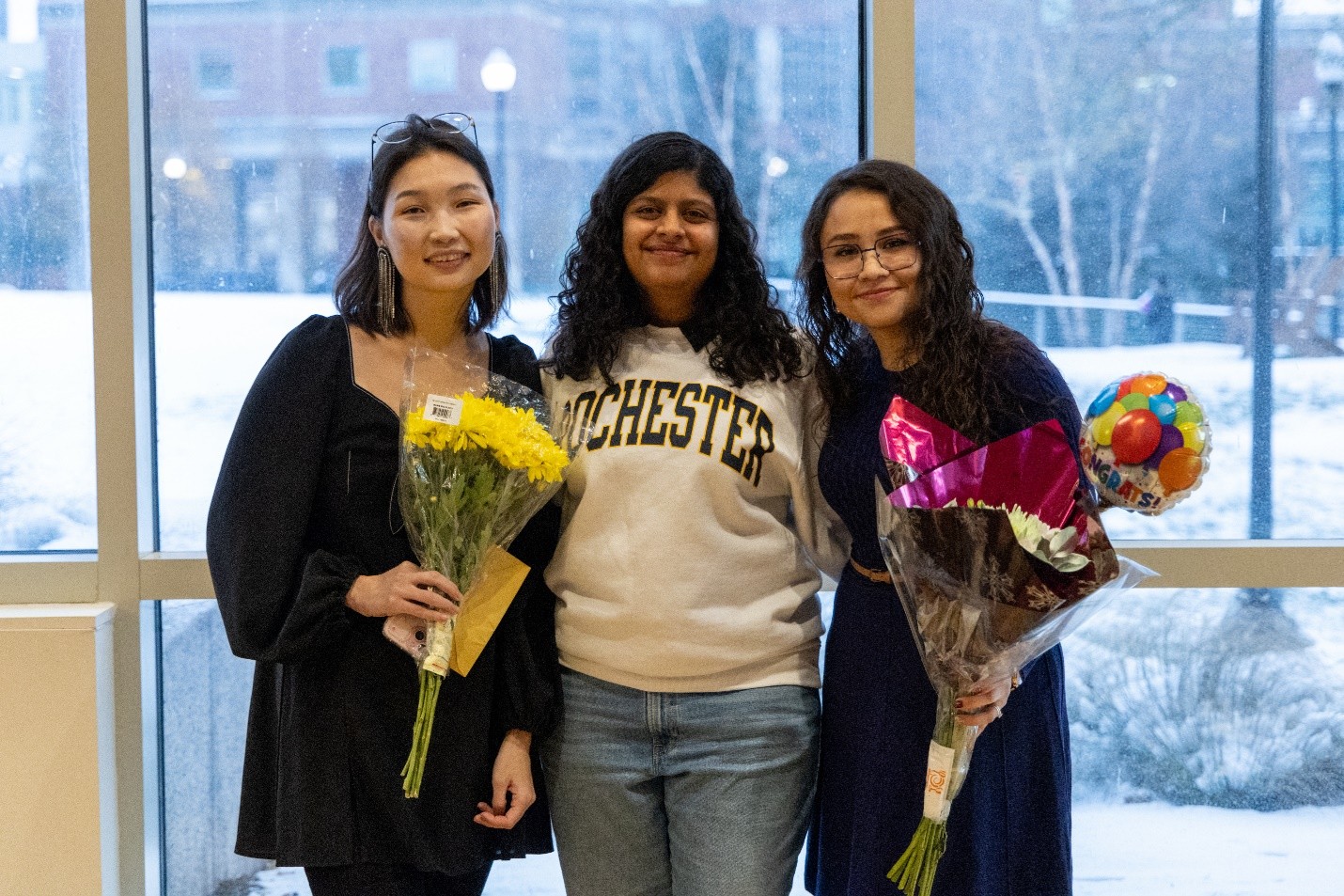 Three people smiling at the camera, two of whom are holding bouquets of flowers.