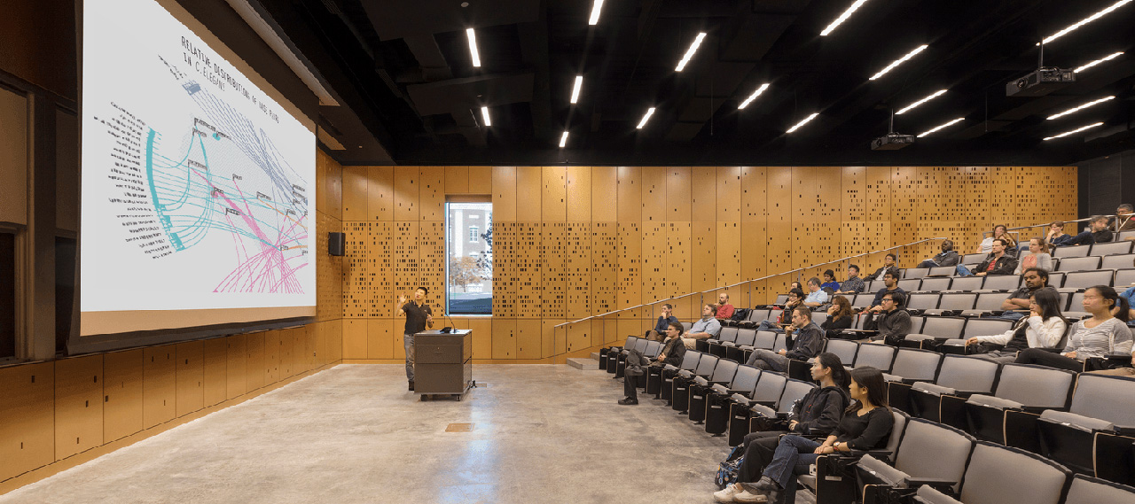 An auditorium-style classroom in Wegmans Hall with a professor behind a podium lecturing to students in seats about a slide on a projection screen.