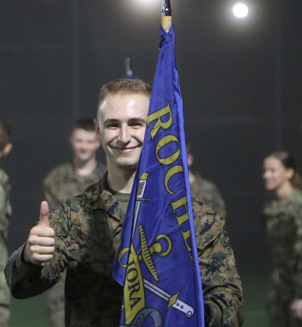 A midshipman holding the UR NROTC flag and giving a thumbs up.