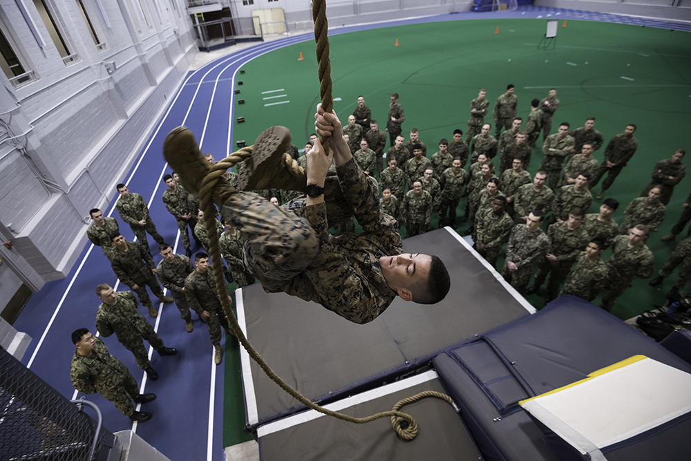 A midshipman climbing a rope while his NROTC class watches from below.