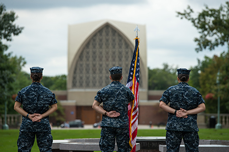 Three midshipman seen from behind standing at attention with an American flag.
