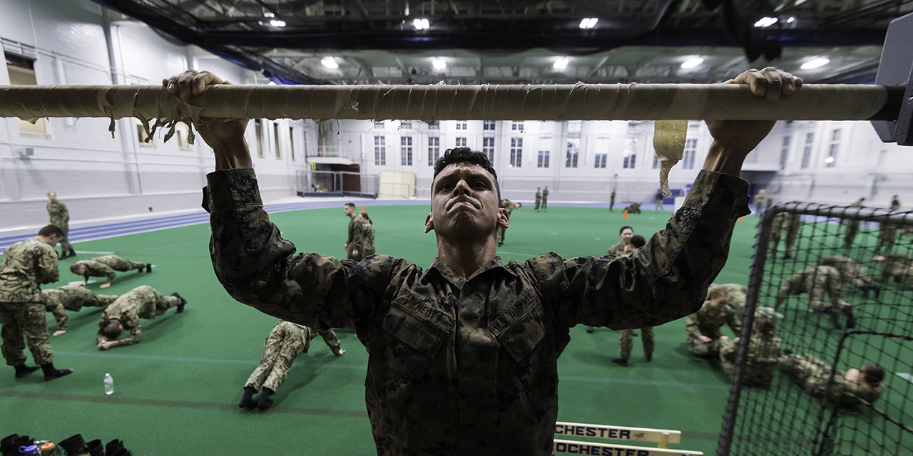 A midshipman doing a pull up while other recruits do exercises in the background.