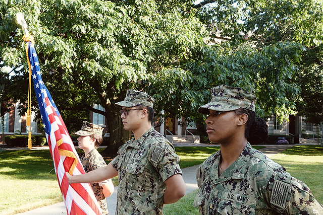 Three midshipman standing at attention with an American flag.