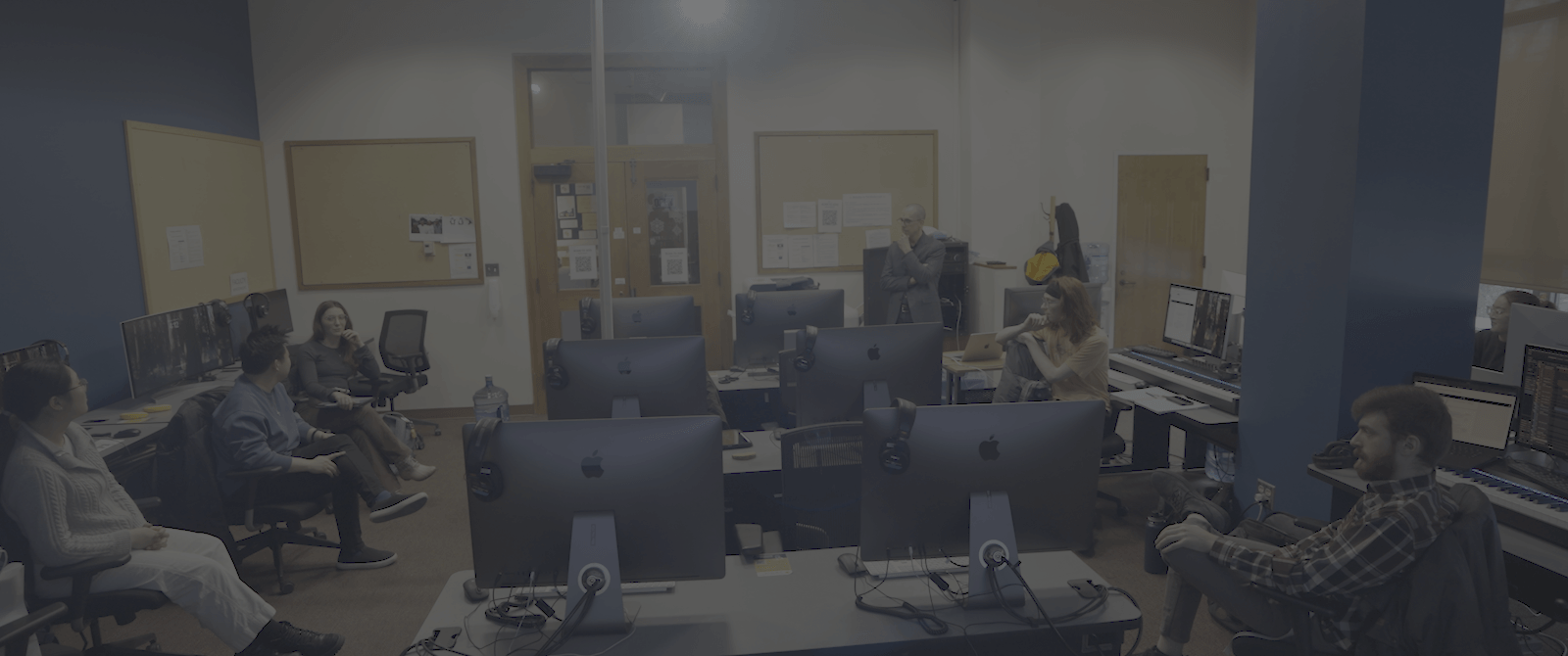 Five students listen to a faculty member while sitting in a lab with keyboards and computers
