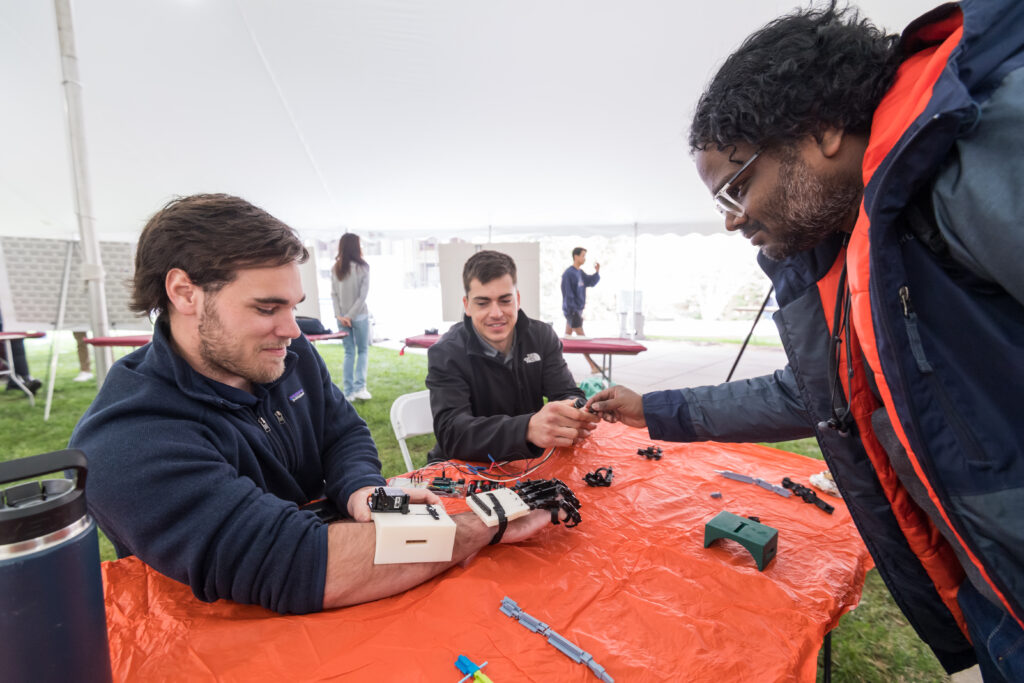 Prashanth Guthikonda watches as URMC Orthopedic Rehab Senior Design Team members Joey Moore (L) and Gabe Lundy model a wearable device that assists the fingers of the human hand to complete its range of motion and return impaired hands to their original strength
